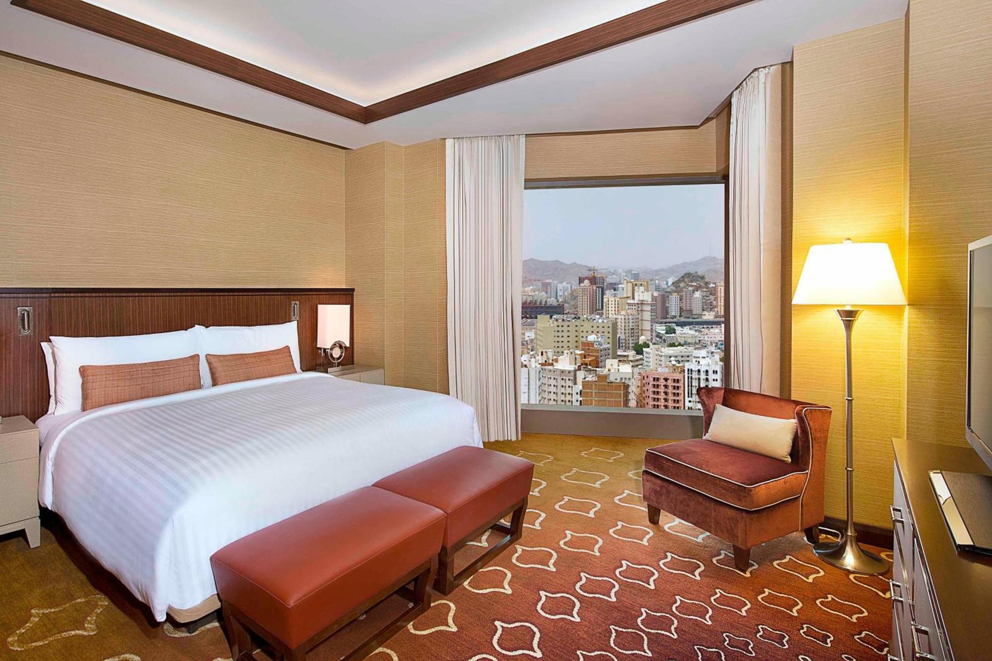 Jabal Omar Marriott Hotel Makkah Mecca Exterior photo The photo shows a modern hotel room featuring a large bed with white linens and decorative pillows. There are two brown ottomans at the foot of the bed, and a comfortable chair next to a window. The room has a warm color palette with light brown wall