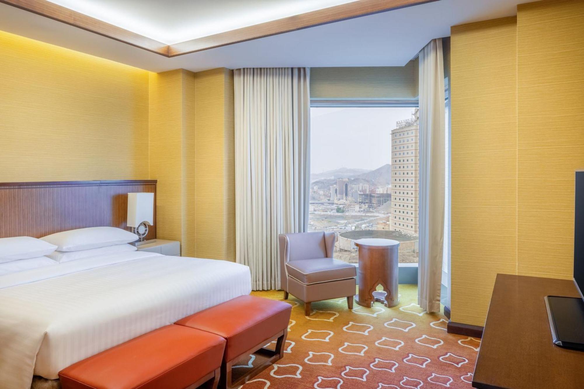 Jabal Omar Marriott Hotel Makkah Mecca Exterior photo The photo shows a modern hotel room with a warm color palette. There is a neatly made bed with white sheets and a brown headboard. In front of the bed, there are two red ottomans. A comfortable chair is placed near a window, which offers a view of th