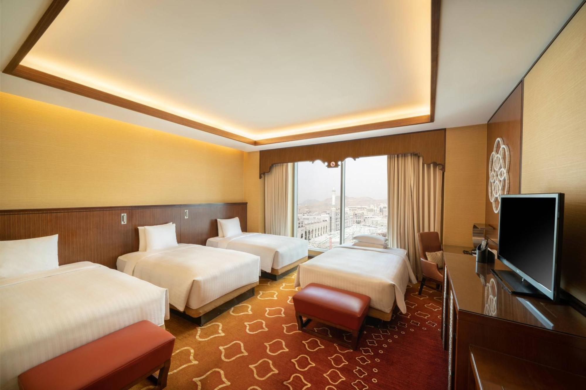Jabal Omar Marriott Hotel Makkah Mecca Exterior photo The photo shows a spacious hotel room featuring four beds arranged in a clean and modern style. The room has a warm color palette with yellow and brown tones. Natural light enters through large windows adorned with curtains, providing a view beyond. 