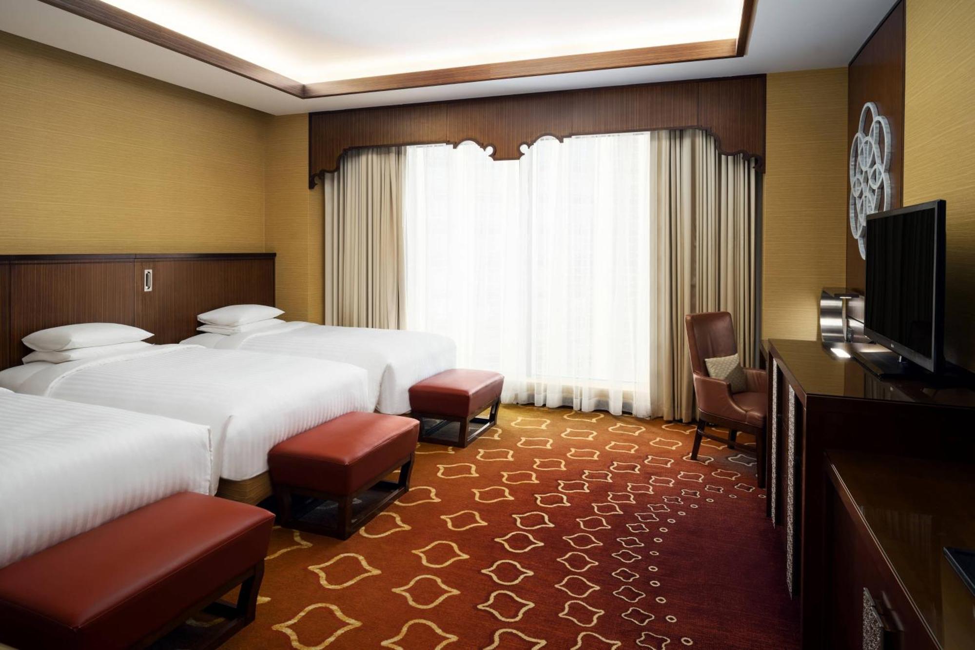 Jabal Omar Marriott Hotel Makkah Mecca Exterior photo The photo shows a well-furnished hotel room featuring three beds with white linens. The room has a warm color scheme, with soft yellow walls and a patterned carpet in shades of red and gold. Large windows with light curtains provide natural light, an